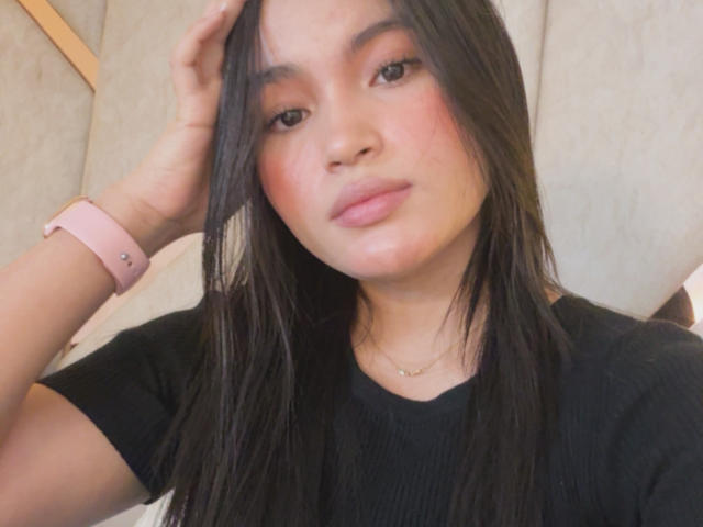 MiiaPalmer met lang zwart haar, een roze horloge, een zwart shirt, lichtrode wangen en roze lippen, kijkt naar de camera, met haar hand in haar haar..