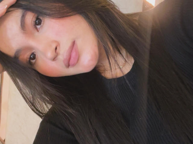 MiiaPalmer with long black hair, wearing a black ribbed shirt, has her hand in her hair, looking at the camera with slightly parted pink lips.