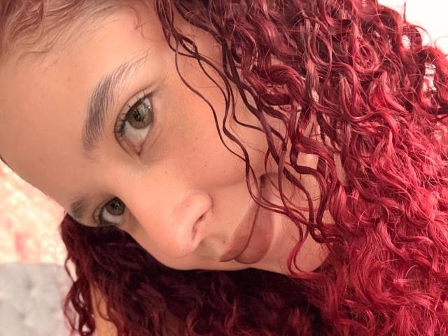 ValeryScot with curly red hair, green eyes, and a pouty face, looking directly at the camera, showing her nose and upper lip, with slightly parted lips.
