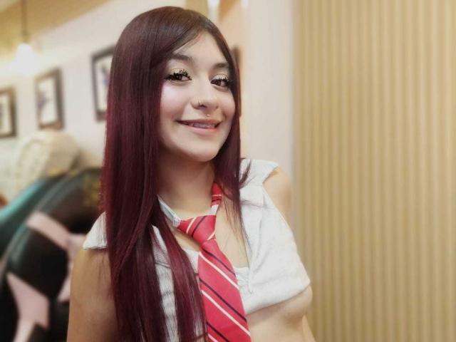 AngelsPhoenix, with long red hair and a cute smile, wears a white shirt and red striped tie, showing off her small tits. She looks happy and relaxed.