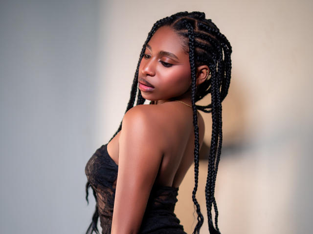 QueenPerlaa with dark skin, long braided hair, looking over her shoulder, wearing a black lace dress, showing her bare shoulders and back, with a subtle, confident expression.