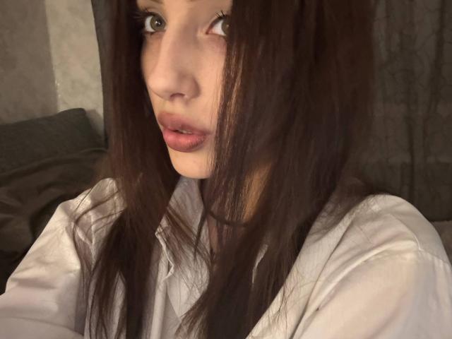 SimonneBrustkern has long brown hair, green eyes, and pink lips. She's wearing a white shirt, looking to the side with a slightly open mouth. Her ass is not visible.