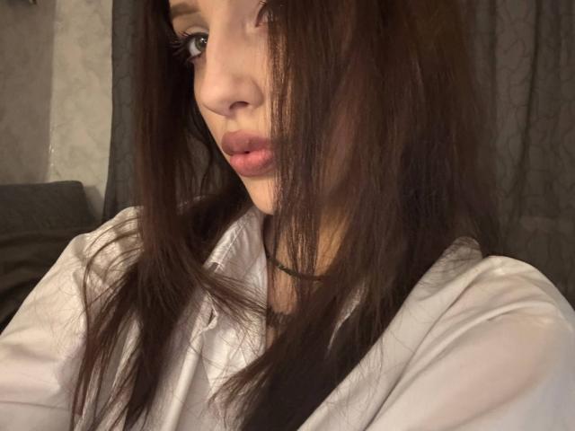 SimonneBrustkern with long brown hair covering her right eye, wearing a white shirt, has pink lips and brown eyes, looking slightly to the side in a soft light.
