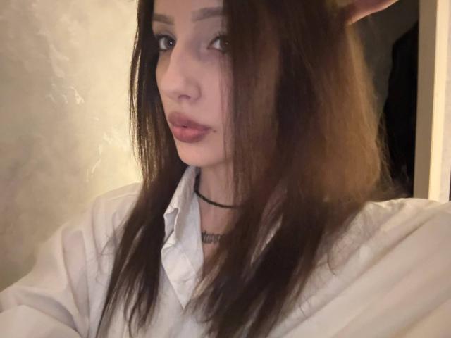 SimonneBrustkern with long brown hair, pointed elf ears, wearing a white shirt and black choker, has pouty pink lips and a slightly flushed face, looking to the side.