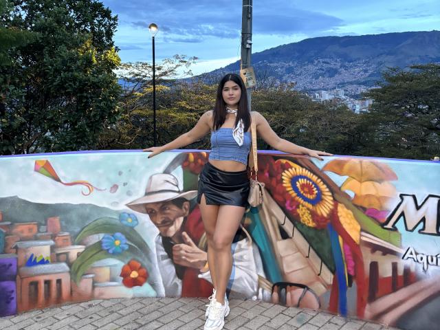 ScarlletZhen, com longos cabelos pretos, vestindo uma blusa cropped azul, uma saia de veludo preta, tênis brancos e uma bolsa branca, está encostada em grafites coloridos. Seus seios são pequenos, e suas nádegas são bonitas..
