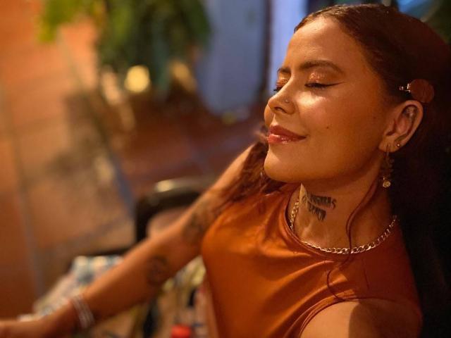 ElectraRoss with brown skin, closed eyes, and a subtle smile, wearing a burnt orange top, has tattoos on her neck and arm, and is sitting in a dimly lit room.