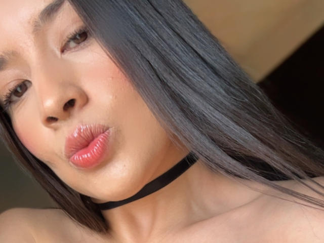 EmmaLombardi with long black hair, pouty pink lips, subtle blush, black choker, and brown eyes, looking at the camera. Her smooth, tan skin and glossy lips stand out.