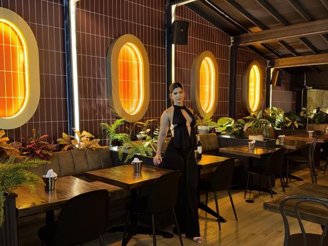 ScarlletZhen está em um restaurante com pouca luz, vestindo um vestido preto de alça, que mostra seus seios e nádegas. Ela tem tatuagens no braço esquerdo e cabelo escuro..