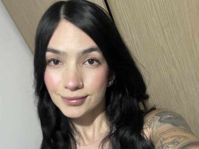 EvaaFarix has black wavy hair, a nose ring, pinkish blush, glossy pink lips, and a tattoo on her right arm. Her brown eyes are slightly upturned.