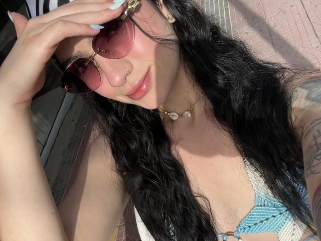 EvaaFarix with pink sunglasses, black wavy hair, pink lips, blue nails, gold necklace, and a blue patterned top, showing a tattoo on her arm, looking at the camera.