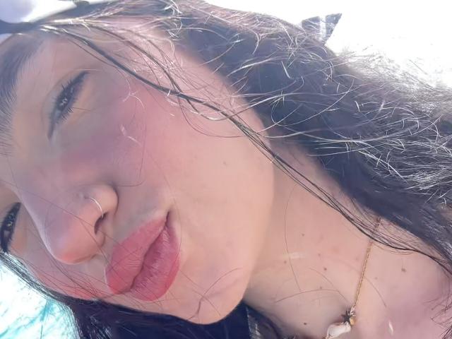 EvaaFarix's puffy pink lips, slightly upturned nose with a small silver nose ring, and long dark hair with strands in her face. She's wearing a gold necklace with a white pendant.