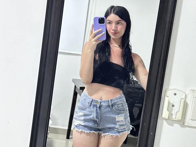 EvaaFarix with long black hair, wearing a black strap top and ripped denim shorts, holding a purple phone, taking a mirror selfie, showing her flat stomach and belly button.