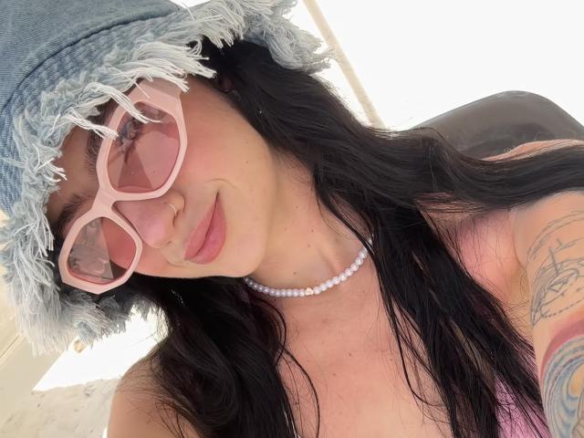 EvaaFarix with pink sunglasses, frayed denim hat, black hair, pink lipstick, pearl necklace, tattoo on right arm, smiling, showing tits, pink tank top, white background.