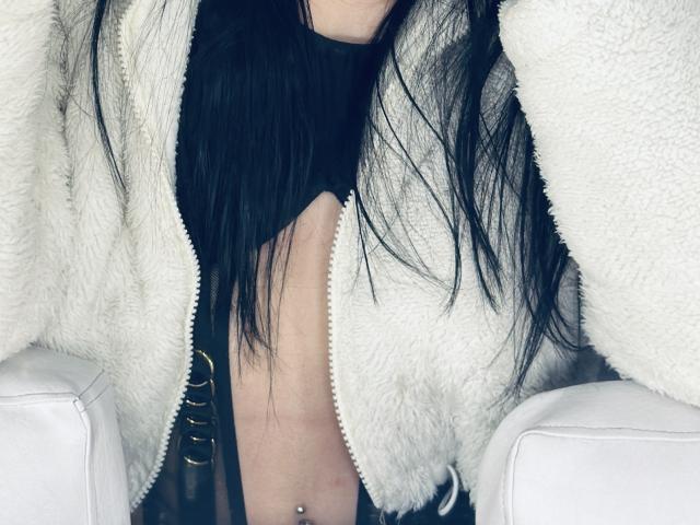 EvaaFarix with long black hair, wearing a fluffy white jacket, exposing her bare chest with a nipple piercing, black top, and gold belt, seated on white furniture.