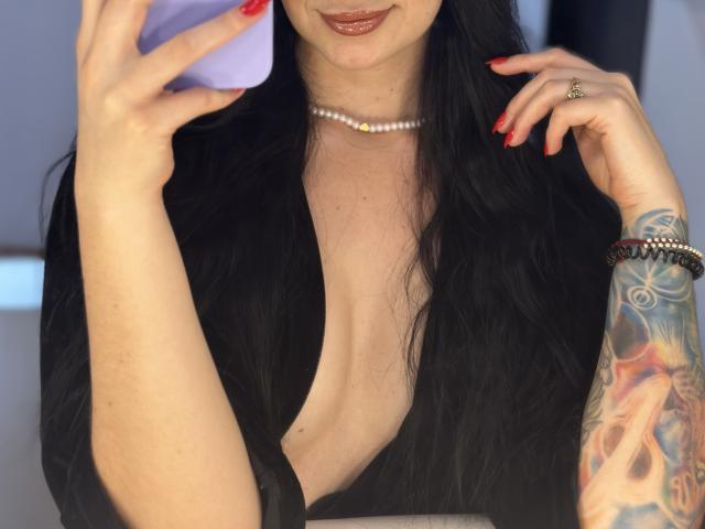 EvaaFarix with black hair, red nails, pearl necklace, deep cleavage, holding phone, visible tattoo on right arm, glossy lips, wearing black top, ring on left hand.