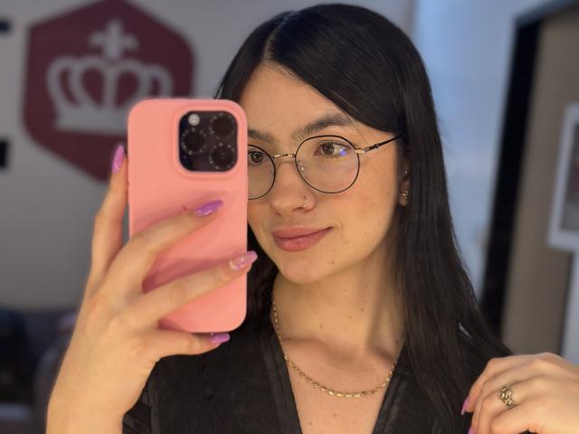 EvaaFarix with long black hair, glasses, pink phone case, purple nails, necklace, and ring, taking a selfie, showing a subtle smile, holding phone in front of face.