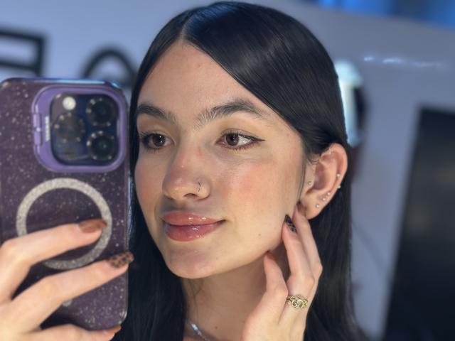 EvaaFarix with glossy lips, holding a phone with a purple case, has black hair, light skin, and a nose ring, with a ring on her finger, smiling softly.