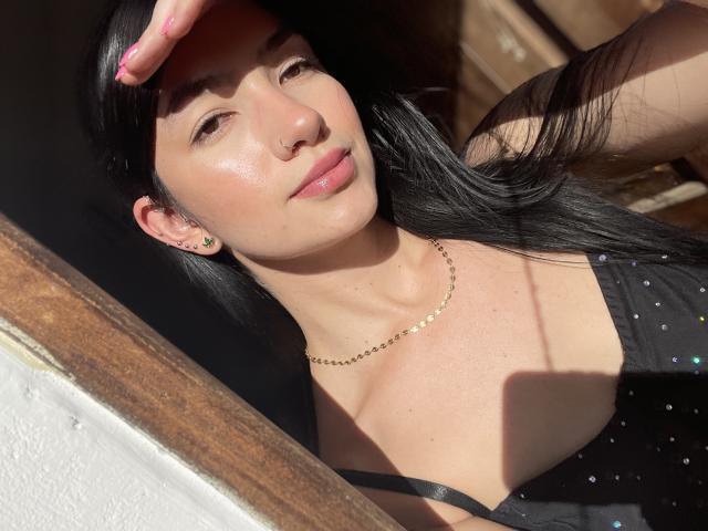 EvaaFarix with long black hair, wearing a black, sparkly top, gold chain necklace, and earrings, looking at camera, sunlight on face, pink nails, slight smile, holding hair up.