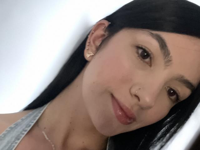EvaaFarix with straight black hair, brown eyes, and a slight smile, wearing a denim top and gold earrings, has a small nose ring and subtle makeup.