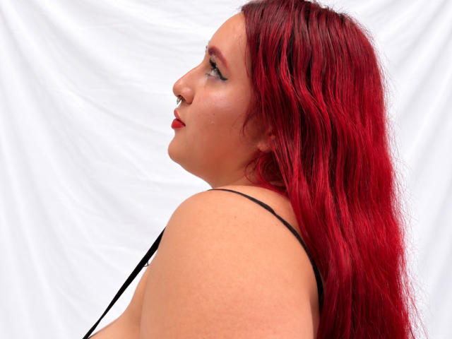 ChoeStar with bright red hair, side profile, wearing a black strap, showing her ass, nose ring, and red lipstick, against a white background.