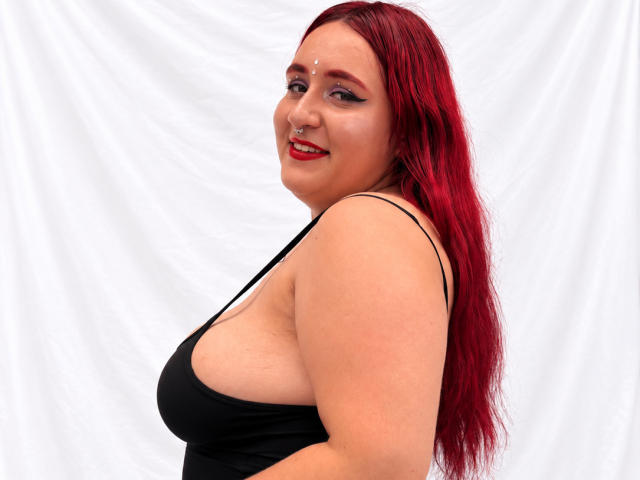 ChoeStar with red hair, black tank top, showing her big tits, smiling, nose ring, red lipstick, white background, standing sideways, looking at camera.