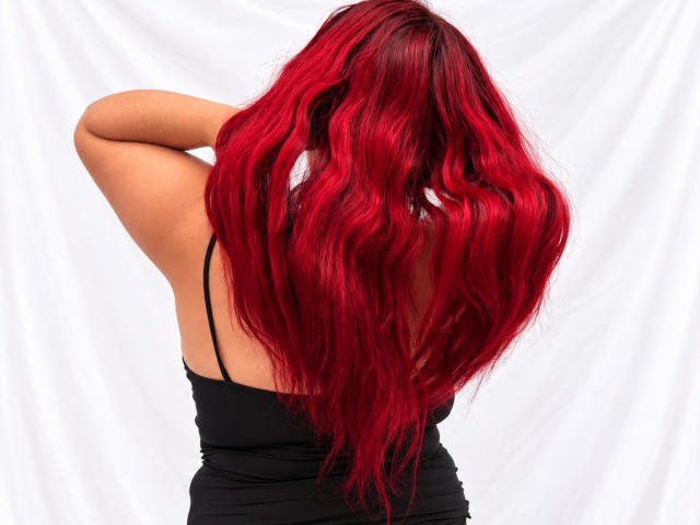 ChoeStar with long, wavy red hair, back to camera, wearing a black spaghetti-strap dress, raising her right arm. Her ass is partially visible.
