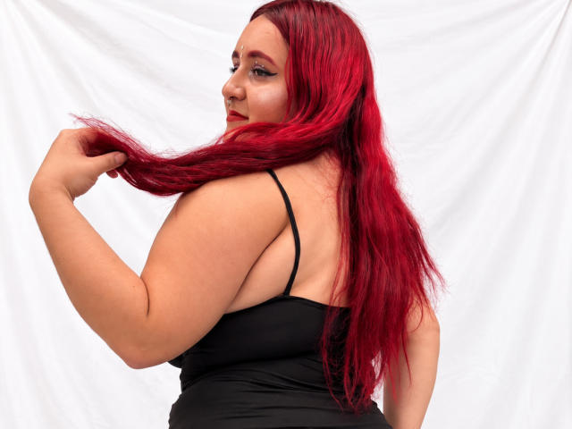 ChoeStar with red hair, holding a long strand, black spaghetti top, looking over shoulder, smiling, fair skin, visible ass, white backdrop, confident, sexy, curvy.