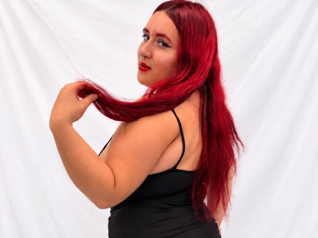 ChoeStar with red hair, holding her long hair, wearing a black spaghetti-strap top, showing her tits, fair skin, red lipstick, and a nose ring, smiling at the camera.
