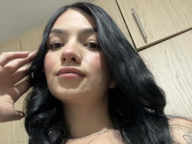 EvaaFarix with black wavy hair, nose ring, and subtle makeup, slightly smiling with a hand in her hair, wearing a necklace, showing a small tattoo on her right shoulder.