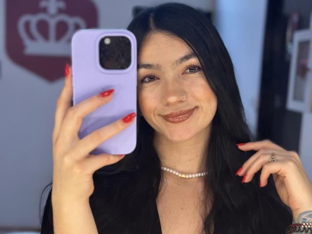 EvaaFarix with black hair, red nails, nose ring, pearl necklace, smiling at phone, holding purple case, taking selfie. Her tits are not visible. Her ass is not visible.