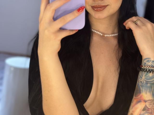 EvaaFarix with long black hair, wearing a black deep V-neck, holding a purple phone, red nails, pearl necklace, and a tattooed arm, showing her small tits and glossy lips.