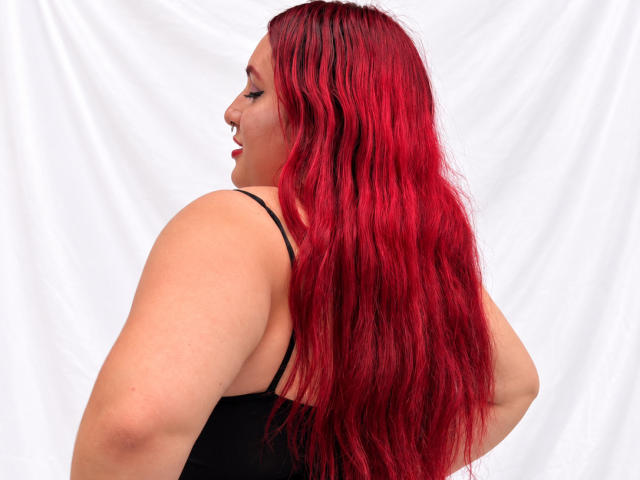 ChoeStar has long, wavy red hair, a fair skin tone, and is wearing a black top. She's looking to the side, showing her back and arms.