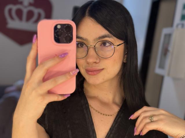 EvaaFarix with glasses, pink phone case, black shirt, pink nails, holding phone, smiling, ring on finger, necklace, dark hair, soft light, nose ring, casual selfie.