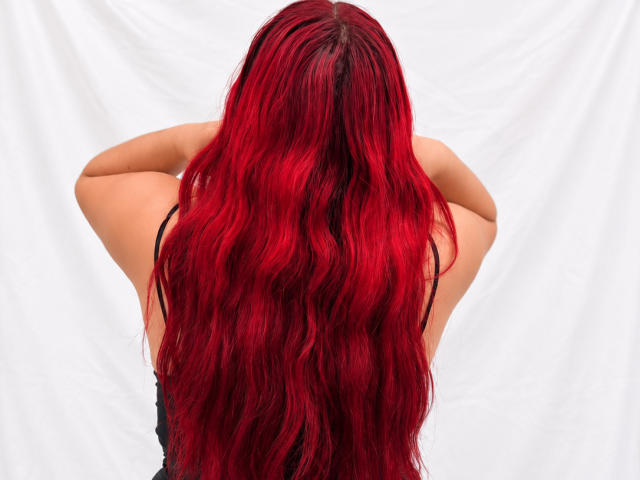 ChoeStar's red, wavy, long hair covers her back, arms raised, wearing a black, strapless top, white fabric backdrop. Boobs partially visible.