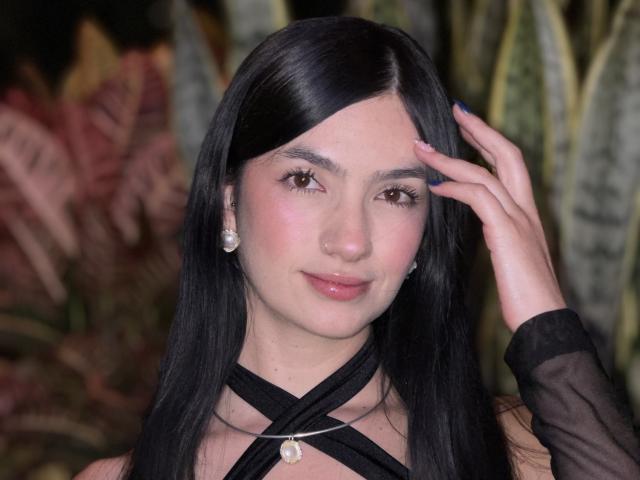 EvaaFarix with black hair, pink blush, and glossy lips, wearing a black criss-cross top with a pearl necklace, touching her hair. She has a cute face and is smiling.