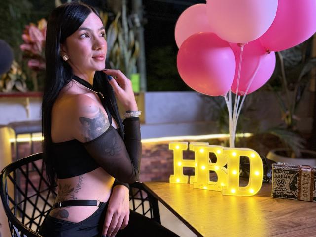 EvaaFarix with black hair, tattoos on arm and side, wearing black top and pants, sitting by table with pink balloons, glowing "HB" sign, wooden table, black chair.