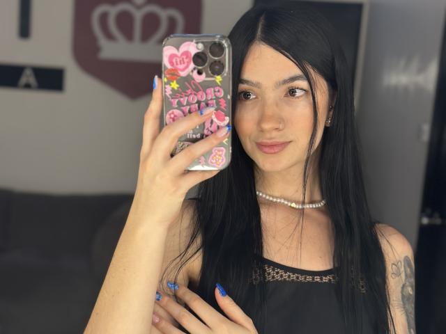 EvaaFarix with long black hair, holding a phone with pink stickers, wearing a black dress, pearl necklace, and blue nails, smiling at the camera, showing her tits.