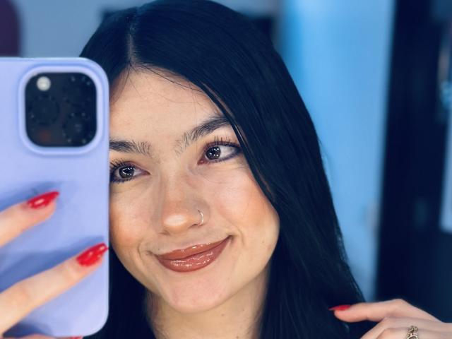EvaaFarix with black hair, red nails, and nose ring, smiling at a phone with blue case, showing shiny brown lips and brown eyes, holding phone in front.