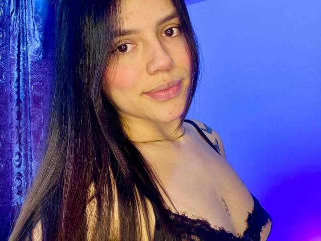 SophiaMarx with long black hair, brown eyes, and a slight smile, wearing a black lace bra, showing her small tits and a tattoo on her right shoulder.
