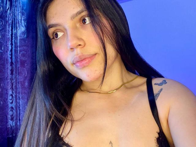 SophiaMarx with long black hair, brown eyes, light skin, small nose, wearing a black lace bra, gold necklace, and tattoos on her shoulder and chest, smiling slightly.