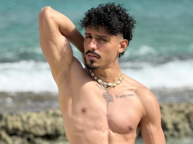 LukeeTaylor, sin camisa, con cabello negro rizado, bigote y collar, levanta el brazo, mostrando el vello axilar, tatuajes pequeños y un cuerpo delgado, musculoso, bronceado con pezones visibles..