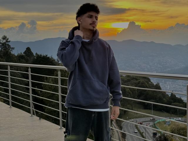LukeeTaylor está em pé, com uma expressão séria, vestindo um moletom cinza e calças verdes, com a mão tocando seu cabelo curto e escuro, ao fundo um pôr do sol com nuvens amarelas e laranjas..