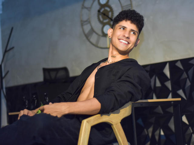 LukeeTaylor, com cabelo preto encaracolado, sorrindo, camisa desabotoada, revelando o peito, calças pretas, cadeira de madeira, segurando uma garrafa verde, relógio ao fundo, pele clara, brincos, casual, relaxada, atraente..