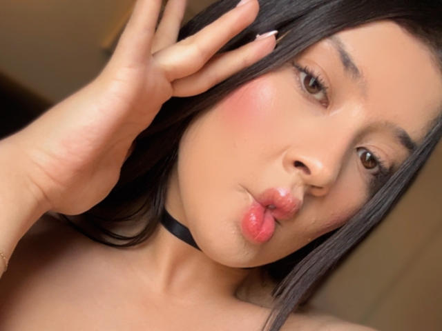 EmmaLombardi has pouty pink lips, blushed cheeks, and black choker. She's touching her face with one hand, brown eyes looking at camera. Her long black hair is in front.