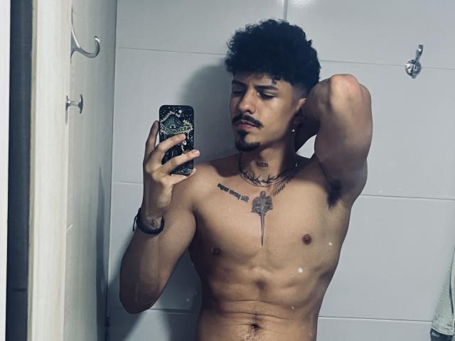 LukeeTaylor, torse nu, avec des cheveux noirs bouclés, tenant un téléphone dans un miroir, avec des tatouages sur la poitrine et le cou, les bras levés, regardant le téléphone, peau brun clair, légère barbe, salle de bain, une serviette visible..