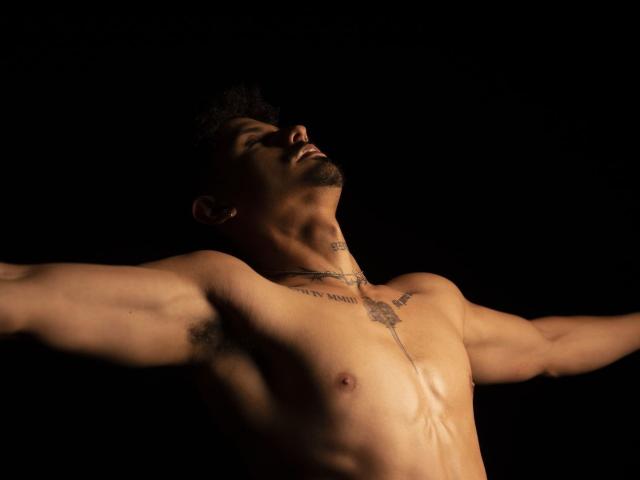 LukeeTaylor, shirtless with arms spread, muscular chest, tattoos on neck, dark short hair, shadowed face, and visible armpit hair, against a black background.