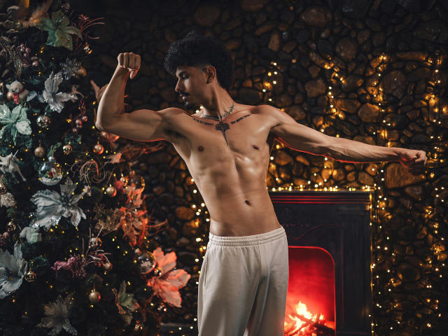 LukeeTaylor, sem camisa, exibindo os bíceps, calças brancas, cabelo cacheado escuro, tatuagem no peito, em pé em frente a uma lareira acesa, árvore de Natal com enfeites à esquerda..