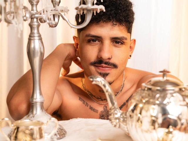 LukeeTaylor, con capelli neri ricci, baffi, piercing al naso e tatuaggi, senza camicia, guarda verso la telecamera. In primo piano, un candelabro d'argento e una teiera. LukeeTaylor indossa una catena d'oro e un orecchino..