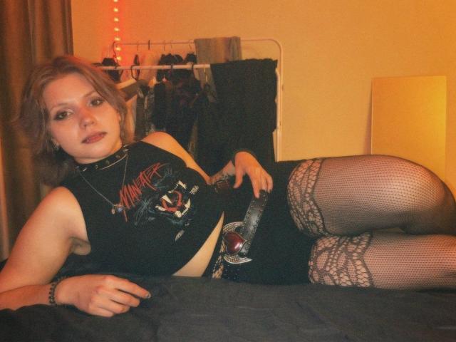 PaolaStohler is lying on a bed, wearing a black top, fishnet stockings with lace, and a belt with a ball gag. She has light brown hair and a neutral expression.