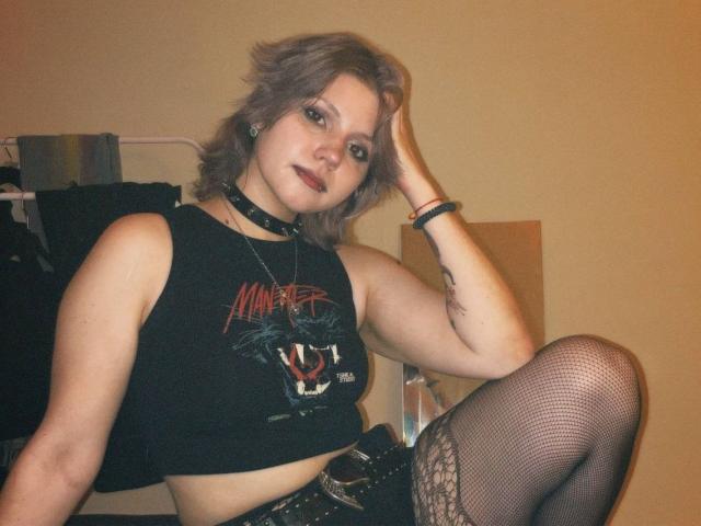 PaolaStohler with short, messy blonde hair, wearing a black "Monster" tank top, fishnet stockings, and a choker, sitting with one leg up, looking at the camera.